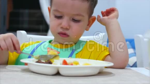 可爱的小孩子坐在围兜里的一张桌子上吃自己的肉泥孩子吃得很心甘情愿可爱的小宝宝吃东西视频的预览图