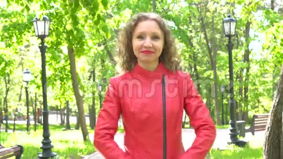 穿着红色皮夹克的年轻女子站在一个美丽的绿色公园里美丽的脸微笑视频的预览图