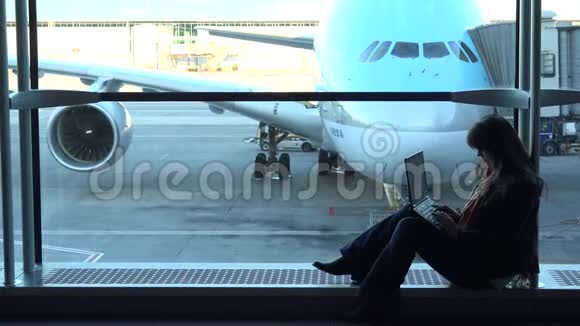 美丽的女人在机场看着笔记本电脑坐在大窗户上飞机停在那里视频的预览图