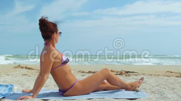 美丽的女人穿着泳衣和太阳镜坐在海滩上看着大海视频的预览图