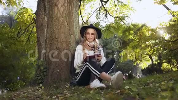 戴着帽子和太阳镜的漂亮年轻女人坐在绿色公园的树旁在她的奶制品里写着东西视频的预览图