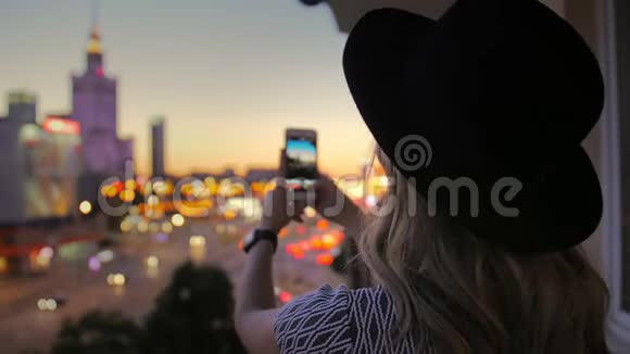 戴帽子的女孩拍了一张夜城的照片使用智能手机在市中心的摩天大楼的框架里视频的预览图