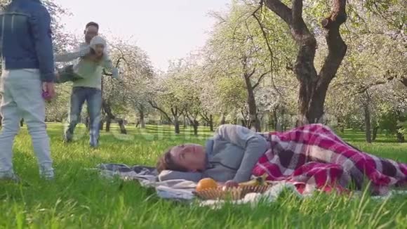 幸福的家庭花时间在盛开的苹果树附近野餐母亲躺在草地上孩子和丈夫跑视频的预览图