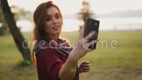 积极的女孩微笑着在智能手机上摆姿势在秋天阳光明媚的公园里自拍视频的预览图