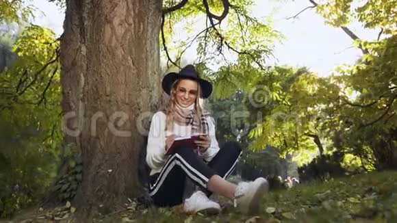 年轻可爱的女人坐在公园背景上的草地上在她的奶制品里做一些笔记视频的预览图