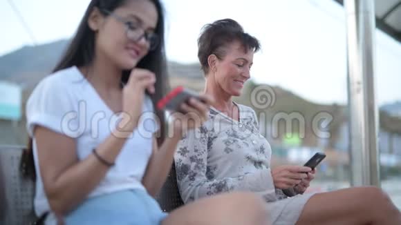 成熟的旅游妇女在与年轻的亚洲旅游妇女等待火车时使用电话视频的预览图