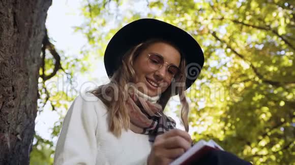 英俊的年轻女子戴着时尚的太阳镜坐在绿色公园里在她的奶制品里写着一些想法视频的预览图