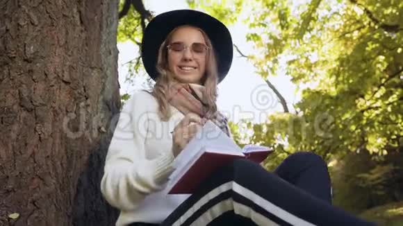 可爱的金发女人戴着帽子坐在公园的树旁在她的笔记簿中注意到了一些想法视频的预览图