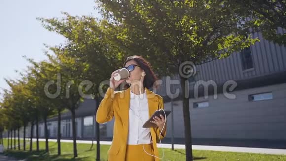 年轻的女商人穿着西装戴着太阳镜手里拿着一台平板电脑在城市街道上喝着咖啡视频的预览图