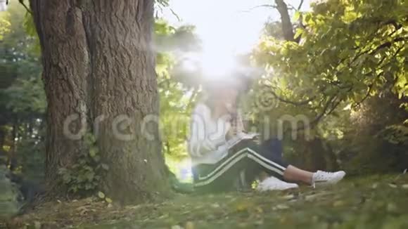 微笑的女人坐在公园的树附近同时在她的笔记本上做一些笔记视频的预览图