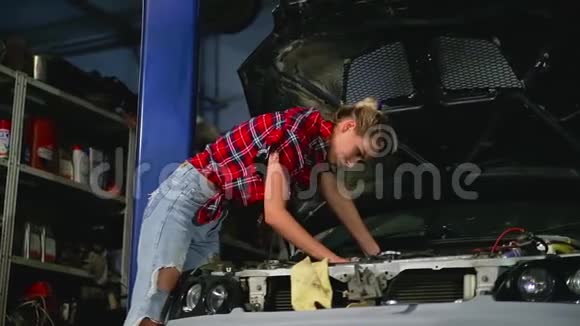 一位穿破牛仔裤的金发女孩在服务站的汽车罩下用扳手拧动发动机的细节视频的预览图