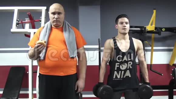 一个胖胖的滑稽男子手里拿着一个谦卑的人站在体育馆里站在年轻英俊的运动员旁边视频的预览图