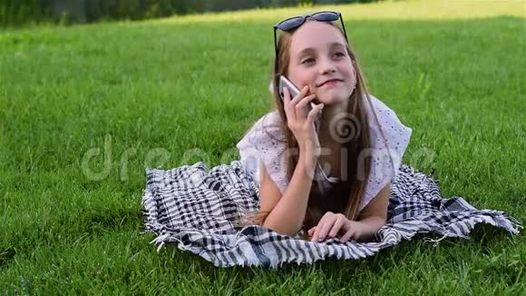 美丽的年轻女孩穿着夏装躺在公园的草地上使用智能手机视频的预览图