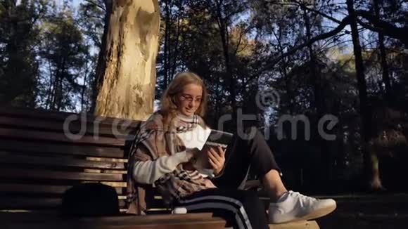 漂亮的年轻女人在绿色公园里坐在长凳上听音乐和使用平板电脑视频的预览图