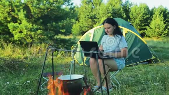 一个年轻的女人坐在帐篷里看着罐子挂在炉火上在笔记本电脑键盘上打字视频的预览图