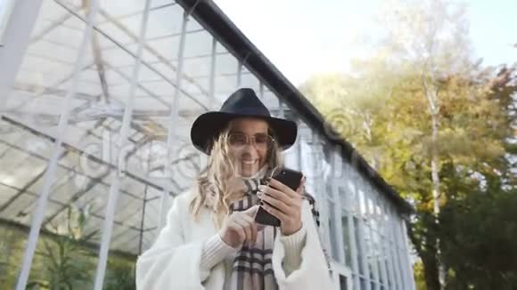 快乐的金发女人戴着太阳镜在公园背景下的智能手机上打字视频的预览图
