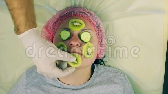 戴手套的化妆师的手从女性脸上取出一个猕猴桃和一根黄瓜的面具视频的预览图