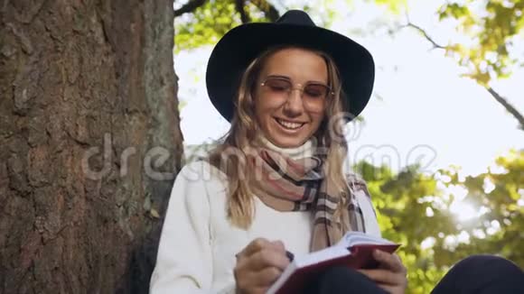 近距离拍摄年轻女子戴着时尚帽子在笔记本上写一些东西同时在城市公园放松视频的预览图