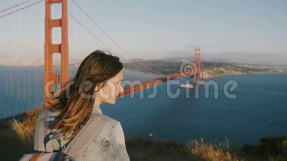 美丽的后景年轻的旅游妇女带着背包头发在风中飘扬欣赏金门大桥的日落视频的预览图