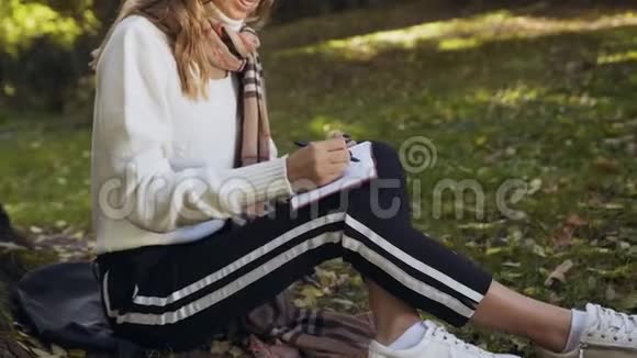 在城市公园里坐在草地上的年轻女子近距离拍摄并在笔记簿上写了一些信息视频的预览图