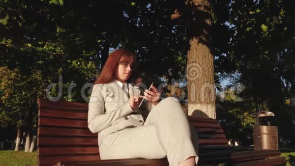 女商人戴着眼镜穿着轻便的西装在夏季公园里的长椅上用平板电脑和电子邮件检查视频的预览图