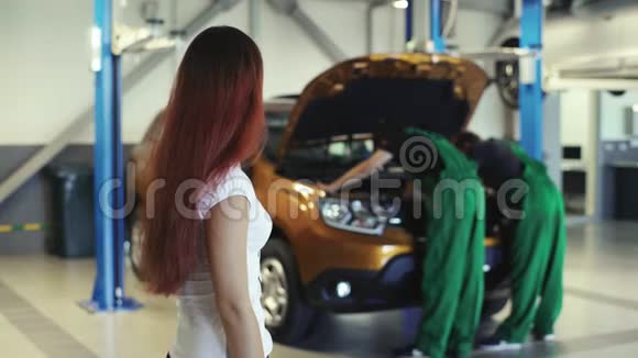 一个漂亮的女孩看着两个机械师在汽车服务中是如何工作的视频的预览图