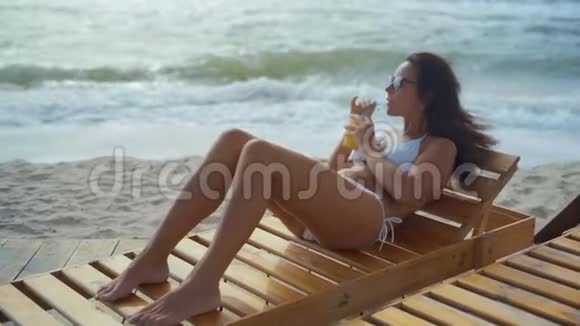 一位穿着白色泳衣和太阳镜的美女躺在海边的木甲板椅上喝着鸡尾酒视频的预览图