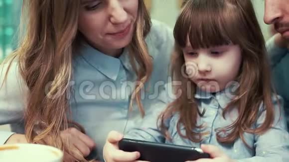 一个小女孩和她的父母在咖啡馆里看着智能手机视频的预览图