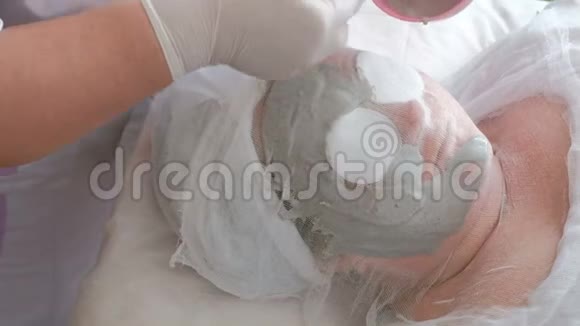 在美容院做皮肤清洁的女人一位白手套的美容师的手在面部涂抹了一团灰色海藻酸钠视频的预览图
