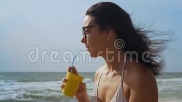 美丽的女人穿着白色泳衣和太阳镜喝着鸡尾酒坐在海边视频的预览图