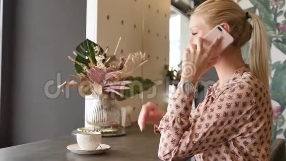 漂亮的金发美女在咖啡馆里用手机说话智能手机女人微笑着在咖啡馆里说话美丽的年轻女性视频的预览图