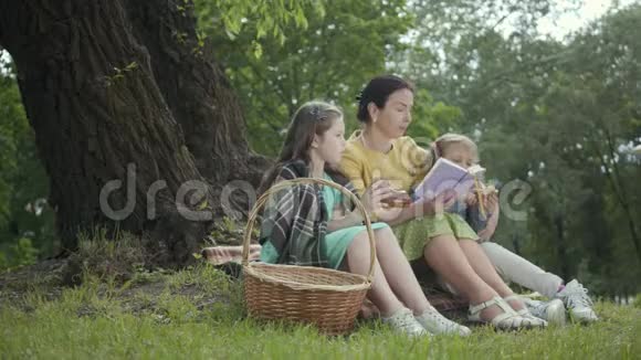优雅的高级女人坐在公园树下的毯子上看书两个可爱的孙女坐着视频的预览图