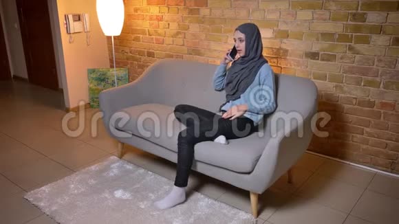 年轻迷人的穆斯林女性戴着头巾坐在舒适的沙发上打电话的特写镜头视频的预览图