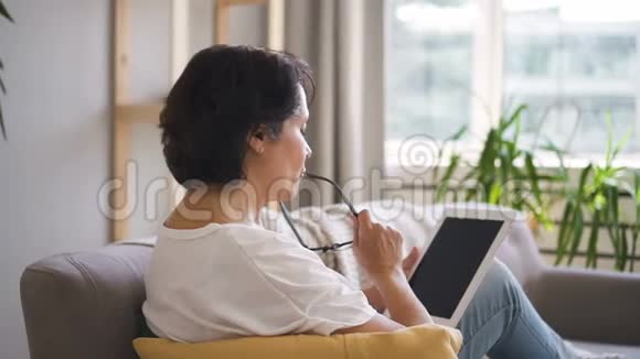 成熟女性的侧视是使用平板电脑坐在家里的沙发上视频的预览图