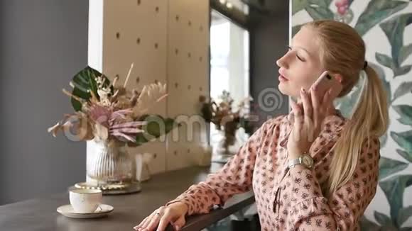 漂亮的金发美女在咖啡馆里用手机说话智能手机女人微笑着在咖啡馆里说话美丽的年轻女性视频的预览图