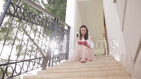 一位拿着数码平板电脑的年轻女子坐在靠近森林的房子前的楼梯上视频的预览图