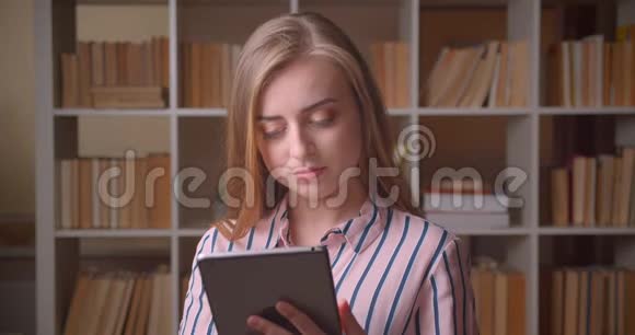 一位年轻漂亮的高加索女学生在大学图书馆里用平板电脑观看照相机的特写照片视频的预览图