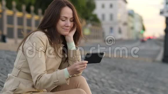 坐在马路附近的城市街道上的女人在智能手机上看书视频的预览图