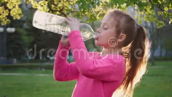 阳光明媚的夏日公园里姜小女孩从瓶子里喝水视频的预览图