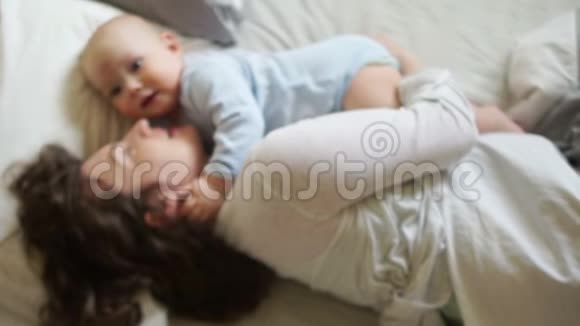 孩子和他妈妈早上躺在床上女人拥抱亲吻婴儿看着镜头微笑视频的预览图