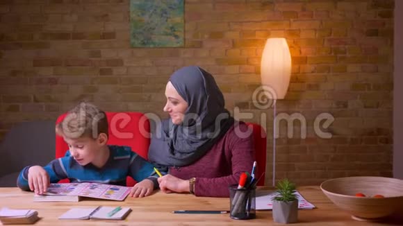 穆斯林母亲戴着头巾和她的小儿子在家里的桌子上学习信件视频的预览图