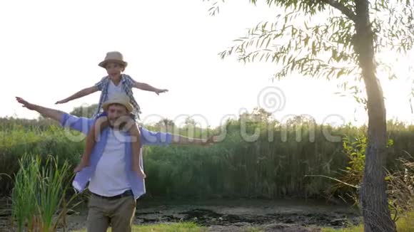 快乐的父亲在背光下和儿子肩上玩耍小男孩想象他在乡下像飞机一样飞行视频的预览图