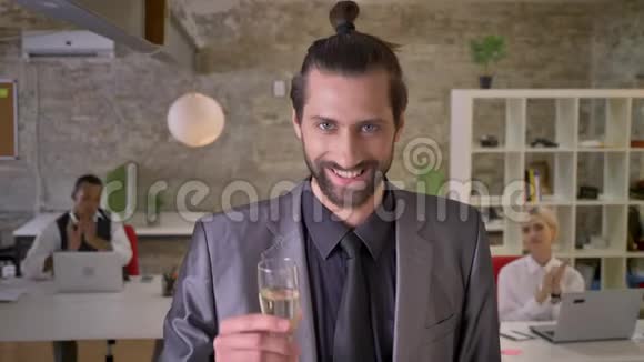 有胡子的有魅力的商人站在办公室里拿着一杯香槟同事们在鼓掌陪伴视频的预览图