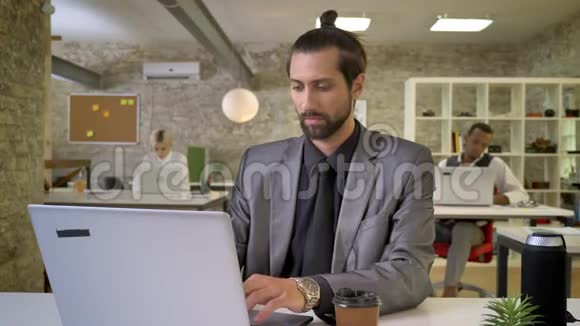 有胡子的漂亮女商人正在办公室里用笔记本电脑打字喝咖啡同事们正在与视频的预览图