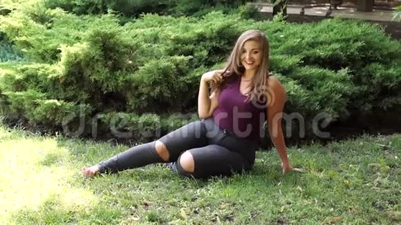 穿着破牛仔裤的胖女孩坐在公园的草地上视频的预览图