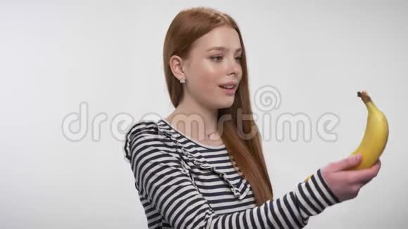 年轻的甜姜姑娘正在剥香蕉咬香蕉享受零食概念白色背景视频的预览图
