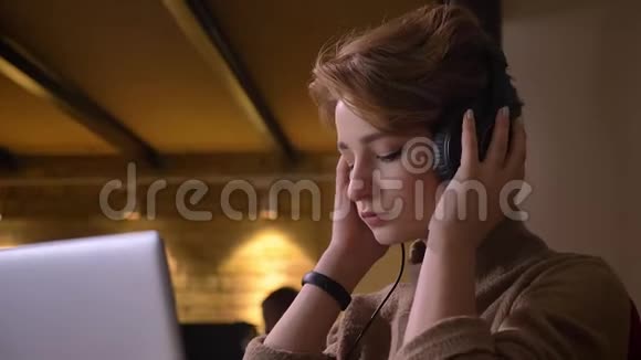 一位年轻的短发女人的特写肖像戴上耳机坐在办公室里听音乐视频的预览图
