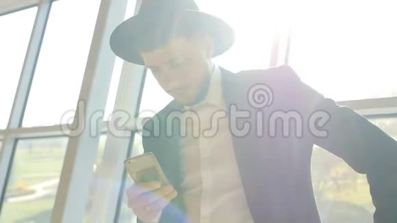 穿着黑色西装戴着帽子的时尚白种人手里拿着电话在机场时看信息视频的预览图