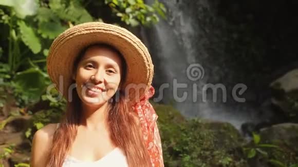 年轻快乐微笑的混合种族旅游女孩的肖像白色的连衣裙和草帽与惊人的野生丛林瀑布生活方式视频的预览图