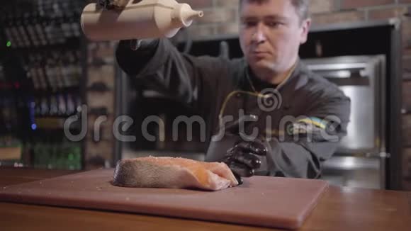 肖像厨师倒入大片三文鱼与糖浆接近穿黑色长袍和橡胶手套的人做菜准备视频的预览图